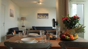 A dining area with a table set for two, a sofa in the background, and a window with a view of a building. - Cosy Apartment in Noordwijk aan Zee With Balcony (Noordwijk)