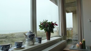 A window with a view of a distant landscape, a teapot, cups, a vase with flowers, and candles on a table. - Cosy Apartment in Noordwijk aan Zee With Balcony (Noordwijk)