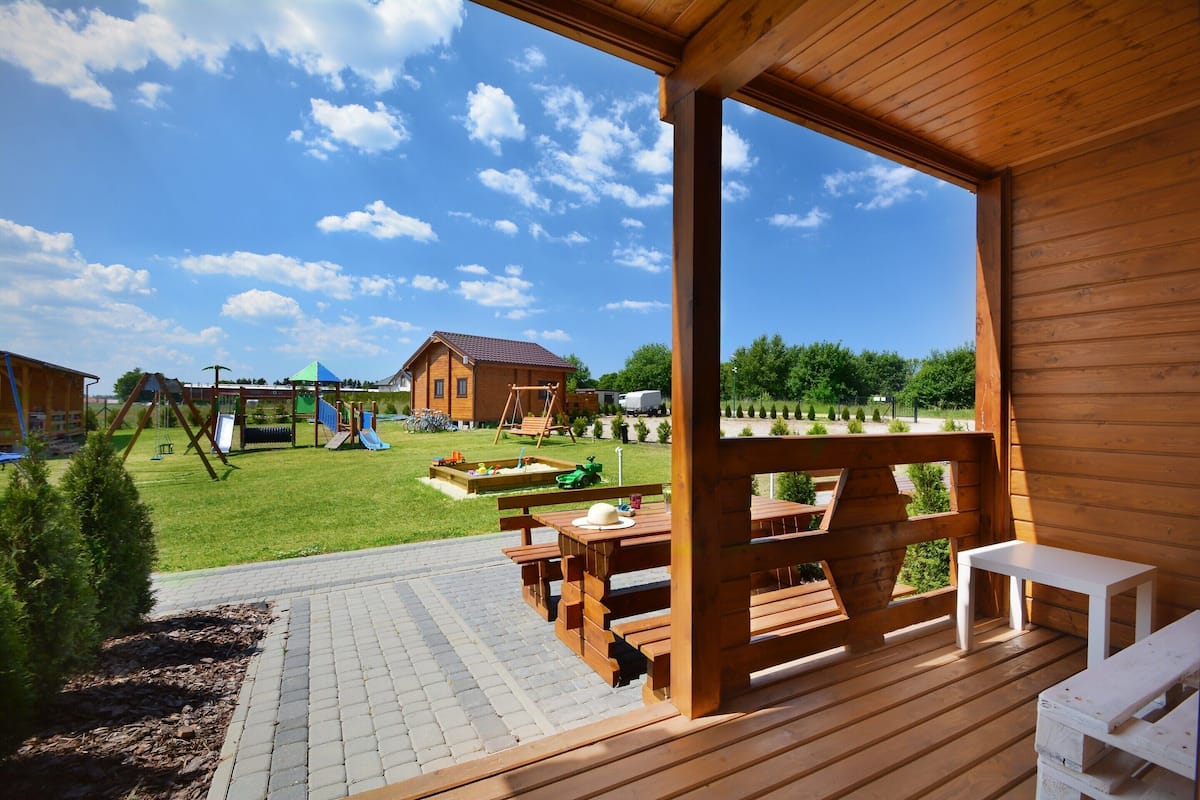 Une terrasse en bois avec des tables de pique-nique, d’où l’on peut admirer une aire de jeux et une maison en arrière-plan.