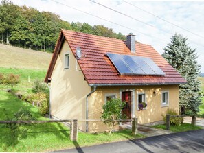 A house with a sloped roof, solar panels, a chimney, and a small porch with a door and windows. - Sachsischer Wald Ruckzug (Hohnstein)