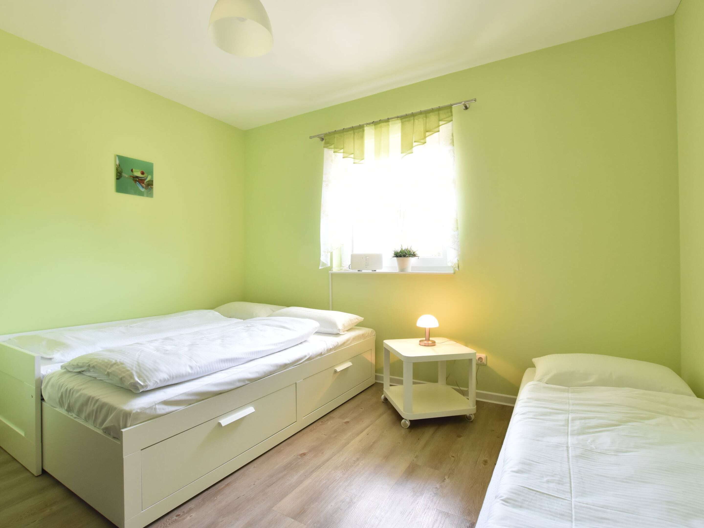 A room with a bed, a bedside table with a lamp, a window with curtains, and a small plant on the windowsill.