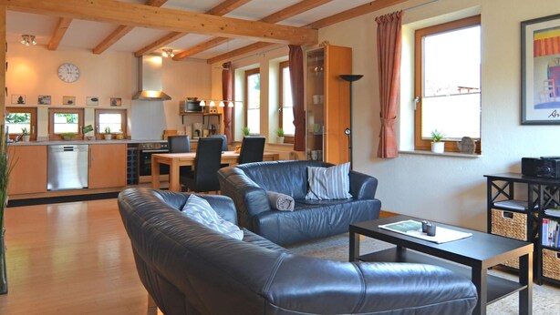 A spacious living area with a sofa, a coffee table, and a dining table. There is a kitchen area with a dishwasher and a microwave. The room has wooden beams on the ceiling and large windows with curtains.
