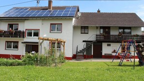 Uma casa de dois andares com painéis solares no telhado, uma pérgola de madeira e um parquinho no quintal.