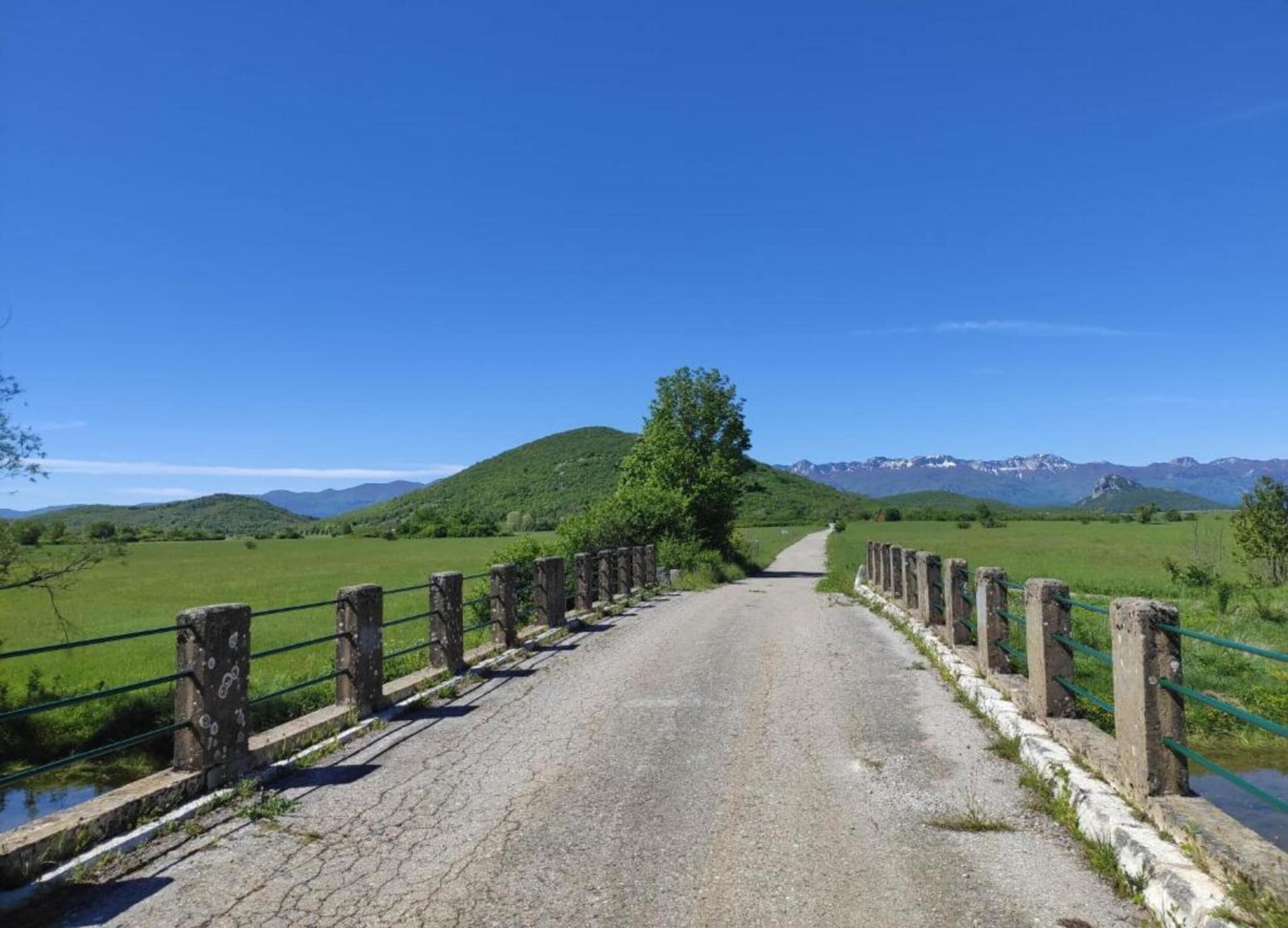 A rural road with stone railings and a clear view of green fields and distant mountains.