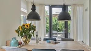 A dining area with a wooden table, four chairs, two pendant lights, a vase with sunflowers, a bottle, and a book. - Beautiful Holiday Home With Terrace (Egmond aan den Hoef)