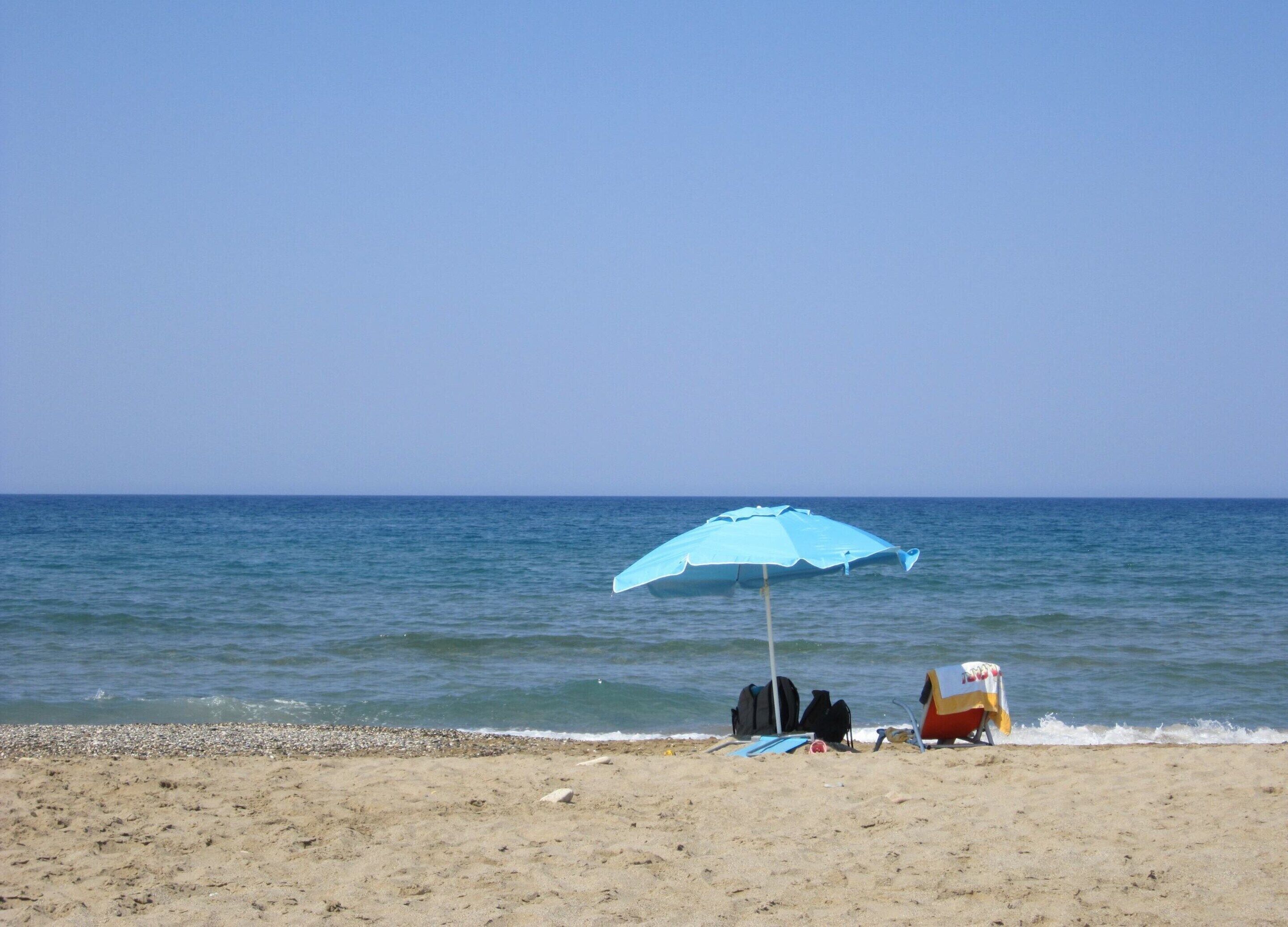 A beach with a blue umbrella, a beach chair, and a small table with a flag.