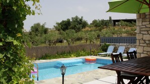 Zona de piscina con una mesa y sillas de madera, una sombrilla verde y un muro de piedra.