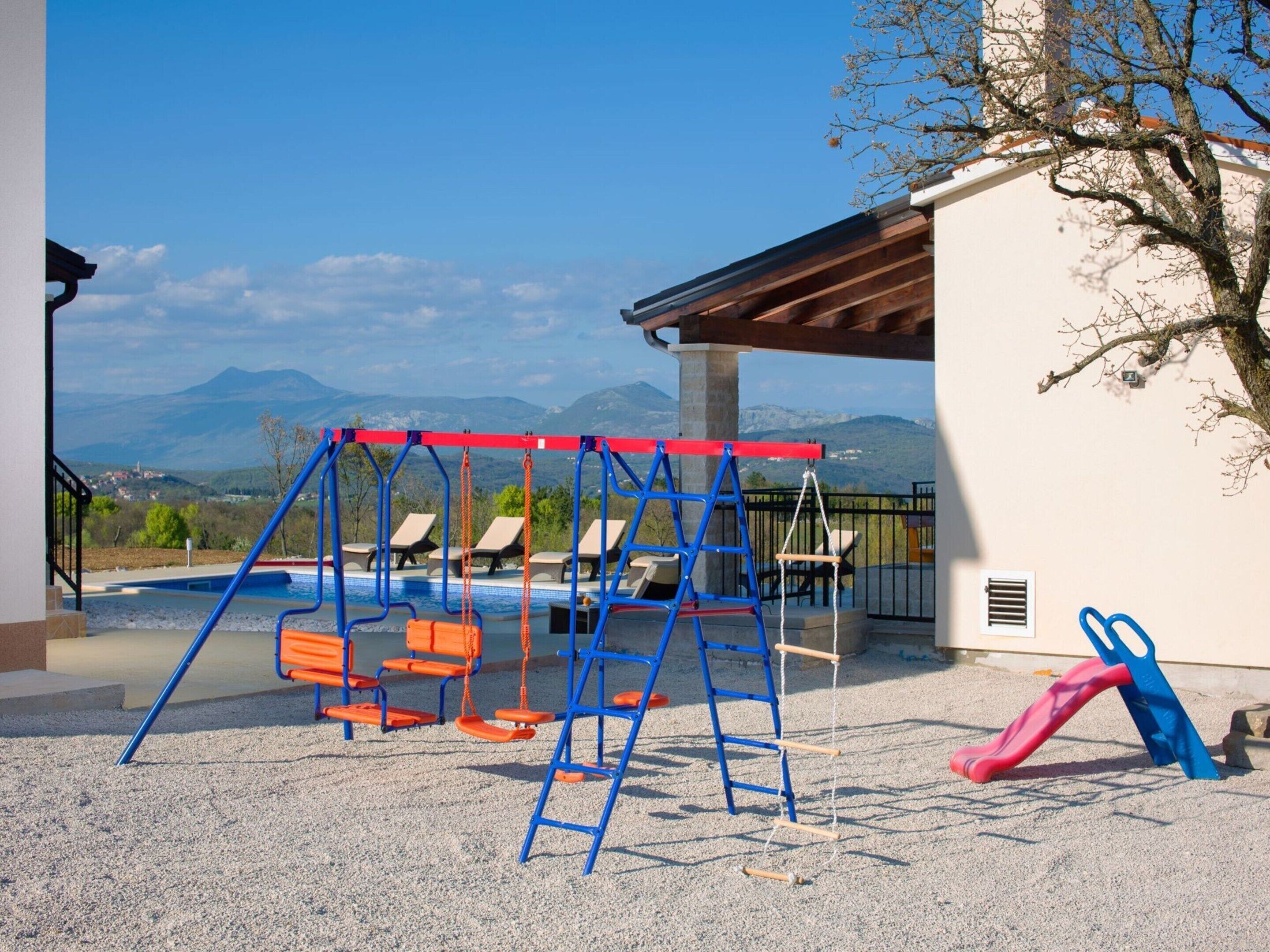 Un parque infantil con columpios y un tobogán, una piscina y vistas a las montañas.