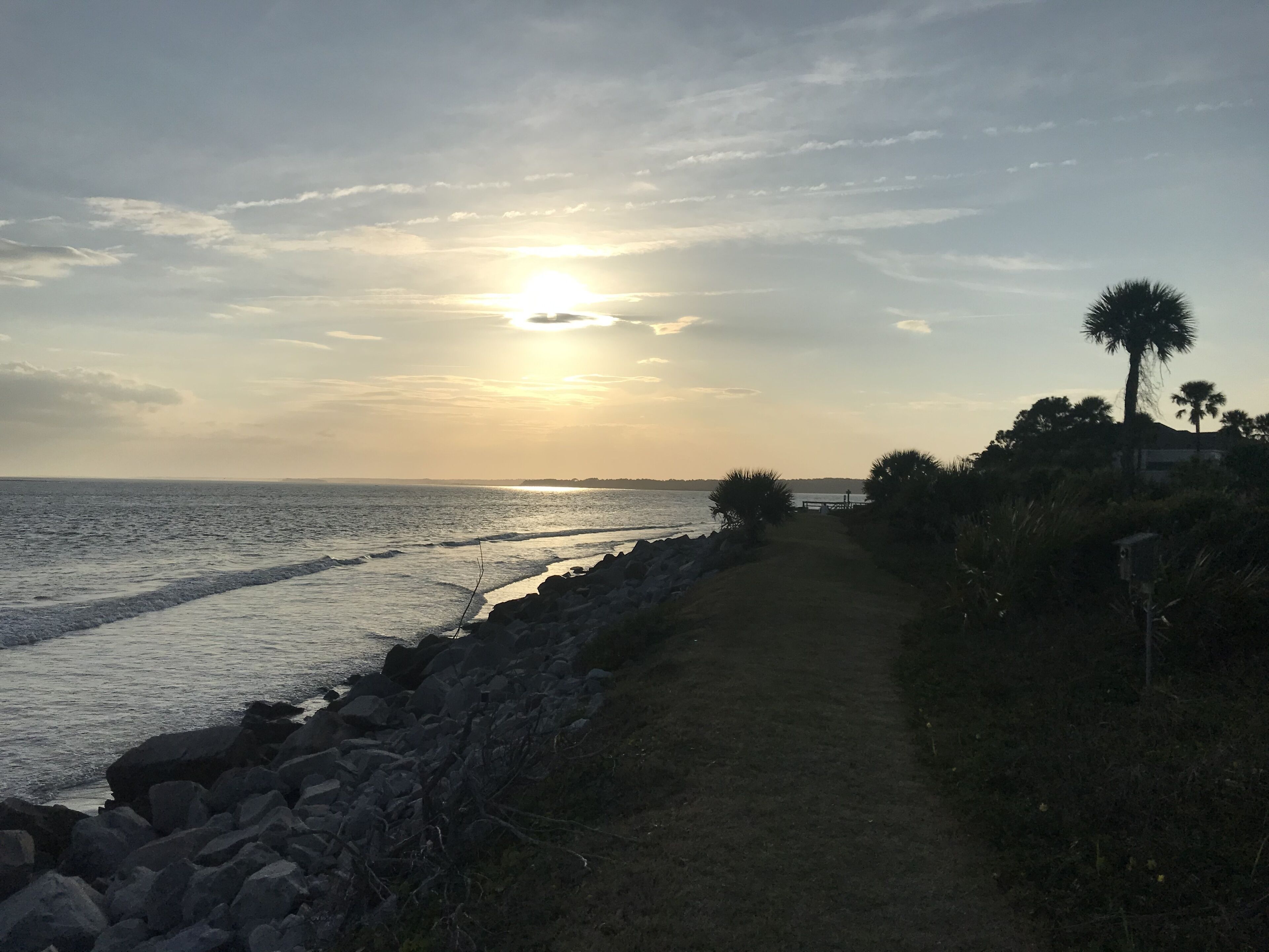 Un sendero costero con un litoral rocoso, palmeras y un atardecer sobre el océano.