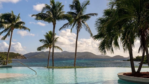Ein Pool mit klarem, blauem Wasser, umgeben von Palmen und Blick auf das Meer und ferne Berge.
