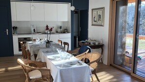 Uma área de jantar moderna com uma mesa posta para quatro pessoas, rodeada por cadeiras de vime. Cozinha com armários brancos e uma mesa de jantar visíveis ao fundo.