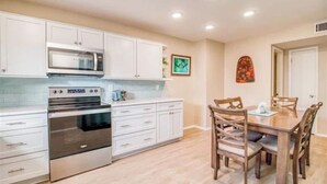 A modern kitchen with white cabinets, stainless steel appliances, and a wooden dining table with chairs. - Entire Pensacola Beach Condo at Boardwalk (Pensacola Beach)