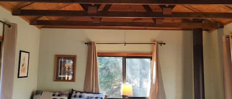 Une pièce chaleureuse avec un plafond en bois, un canapé agrémenté de coussins à motifs, une fenêtre avec des rideaux, une lampe fixée au mur et un tableau encadré.