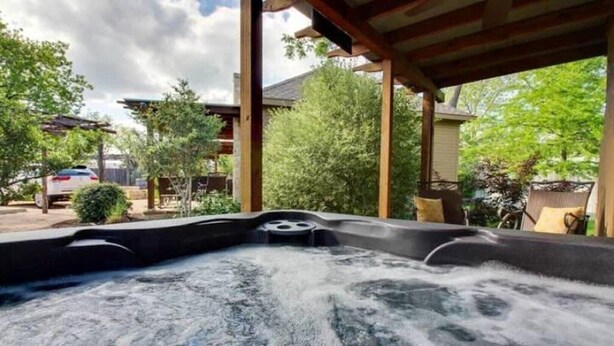 Un bain à remous surmonté d’une pergola en bois, entouré d’arbres et avec une voiture garée à proximité.