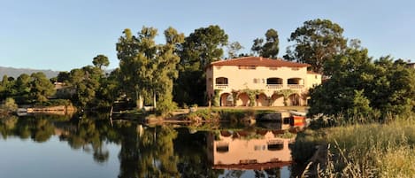 A building with arched windows and a red-tiled roof, surrounded by trees and a calm body of water reflecting its structure.