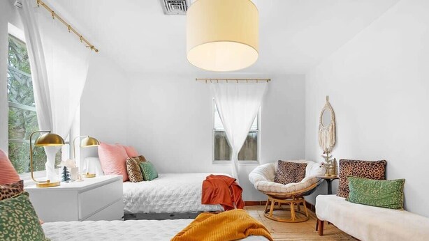 Une chambre avec deux lits, un coin salon avec un fauteuil et un canapé, une grande fenêtre avec des rideaux et un luminaire suspendu.