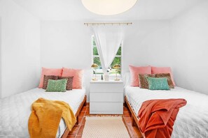 Une chambre avec deux lits, des murs blancs, une fenêtre avec des rideaux et une table de chevet avec une lampe.