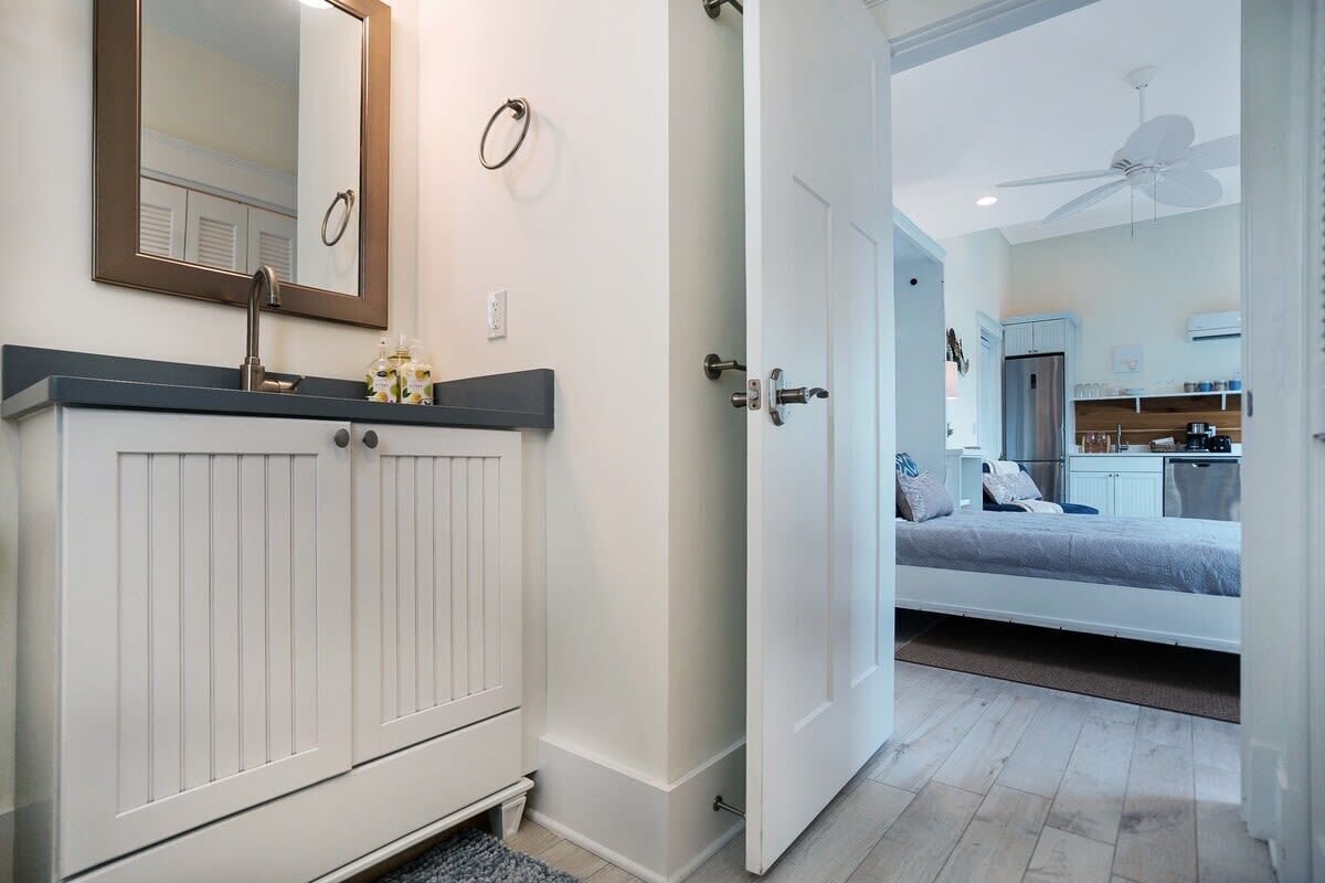 A modern bathroom with a white vanity, a mirror, and a door leading to a bedroom with a bed and a kitchenette.