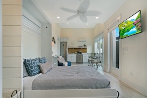 A bedroom with a bed, a ceiling fan, a TV, and a kitchen in the background. - 100 Steps to Beach, Deck w Ocean View, Beach Gear (St. Augustine)