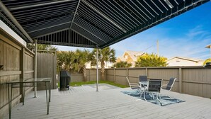 A covered outdoor patio with a table and chairs, a grill, and a wooden deck.