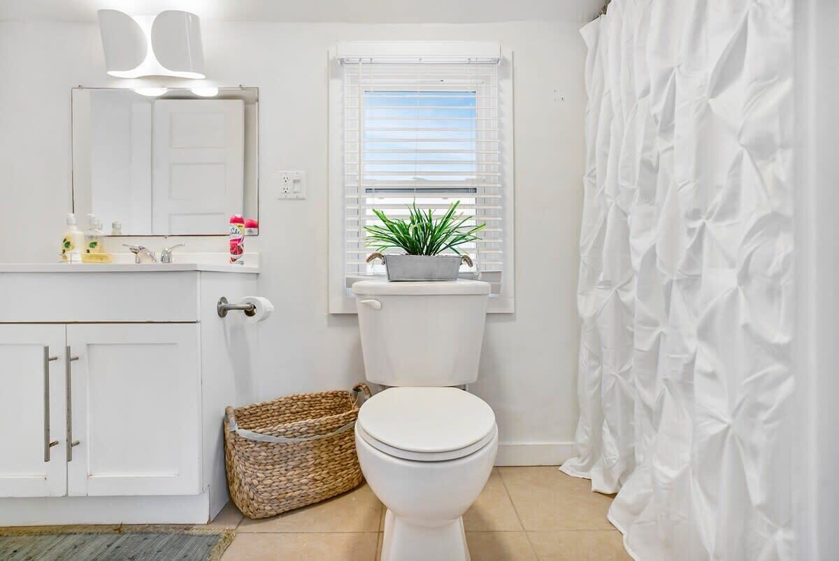 Een badkamer met een wit toilet, een wastafel met toiletartikelen, een spiegel, een raam met zonwering en een douchegordijn.