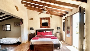 A bedroom with a large bed, wooden beams, a ceiling fan, a small bedside table, a nightstand, a mirror, and a sliding glass door leading to an outdoor area. - Large traditional CORTIJO, Large Private pool, Ping Pong table , family/groups (Malaga)