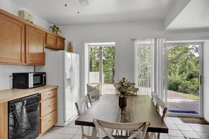 A kitchen with wooden cabinets, a white refrigerator, a microwave, and a dishwasher. There is a dining table with chairs and a vase with flowers. The room has large windows with a view of trees and a house. - The Hideaway -Raystown Lake Vacation Rentals, Aunt Susie's (Hesston)