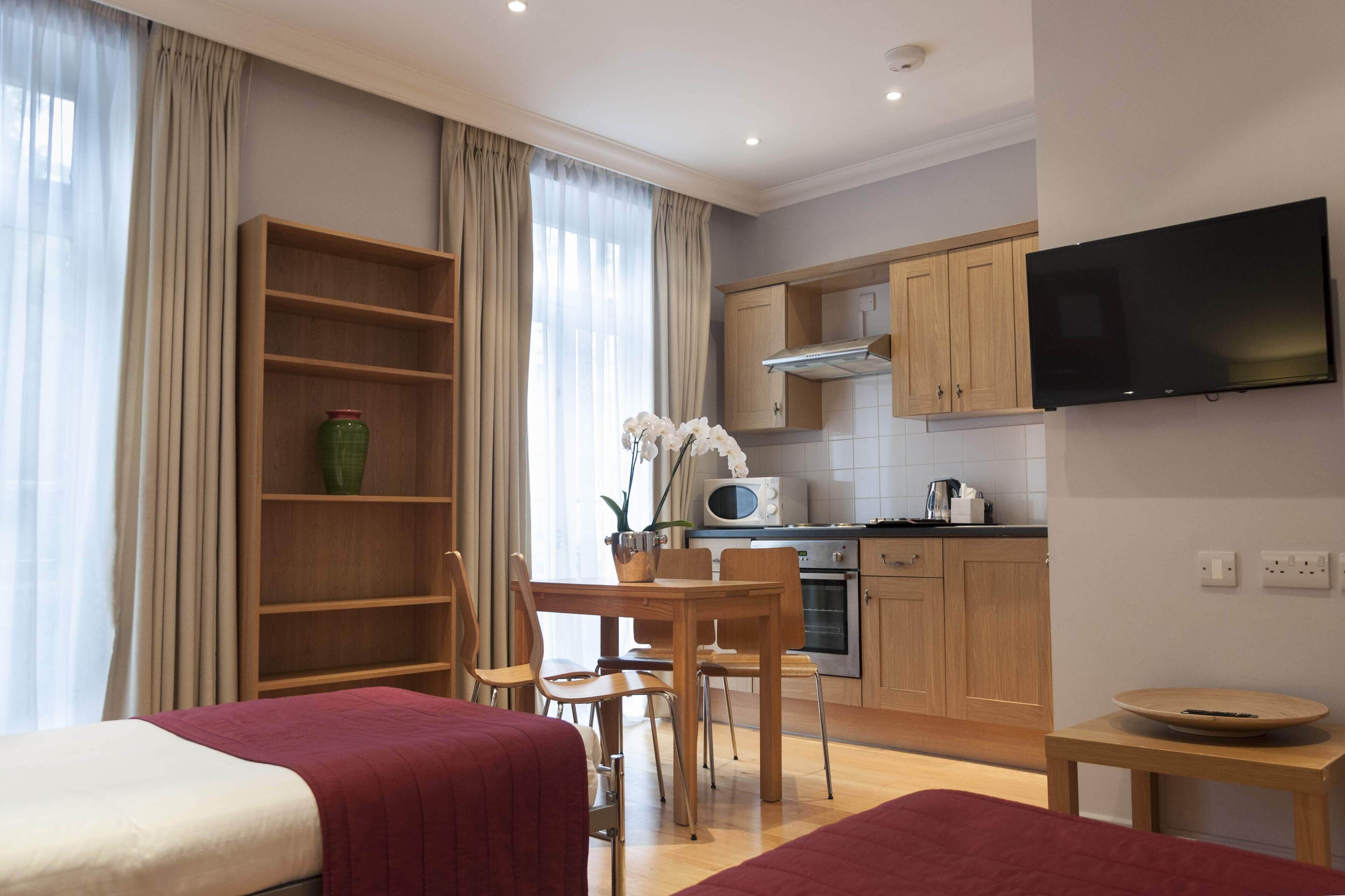 A modern hotel room with a bed, a desk, a chair, a TV, and a kitchenette.