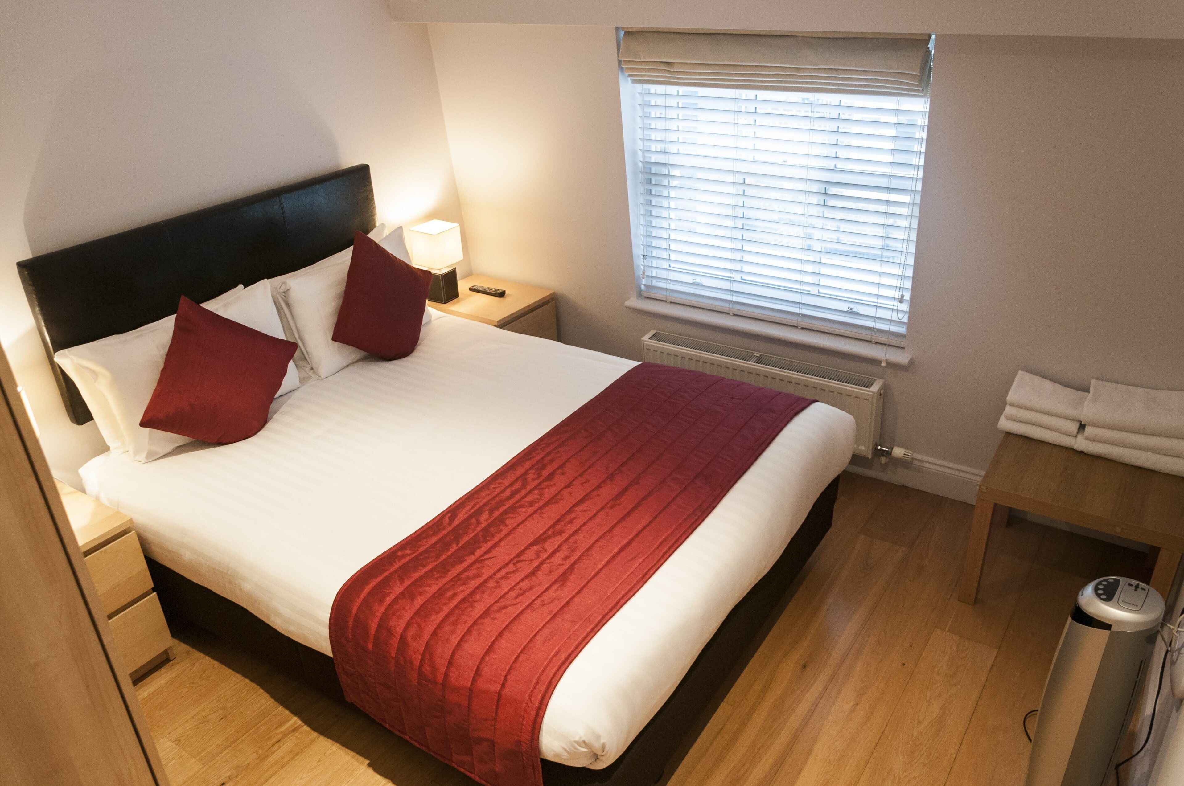 Ein ordentlich bezogenes Bett mit einer roten Decke, ein Nachttisch mit einer Lampe und ein Fenster mit Jalousien.