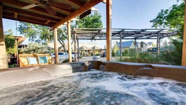 Un área de hidromasaje con una pérgola de madera, rodeada de follaje y vistas a coches aparcados y casas.