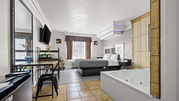 A hotel room with a bed, a desk with a chair, a TV, a large window with curtains, a bathroom with a bathtub, and a tiled floor. - Rodeway Inn Santa Ana - Garden Grove (Santa Ana)