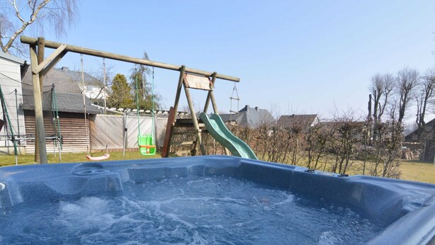 Ett bubbelbad i en trädgård, med en gungställning i trä och en grön rutschkana.