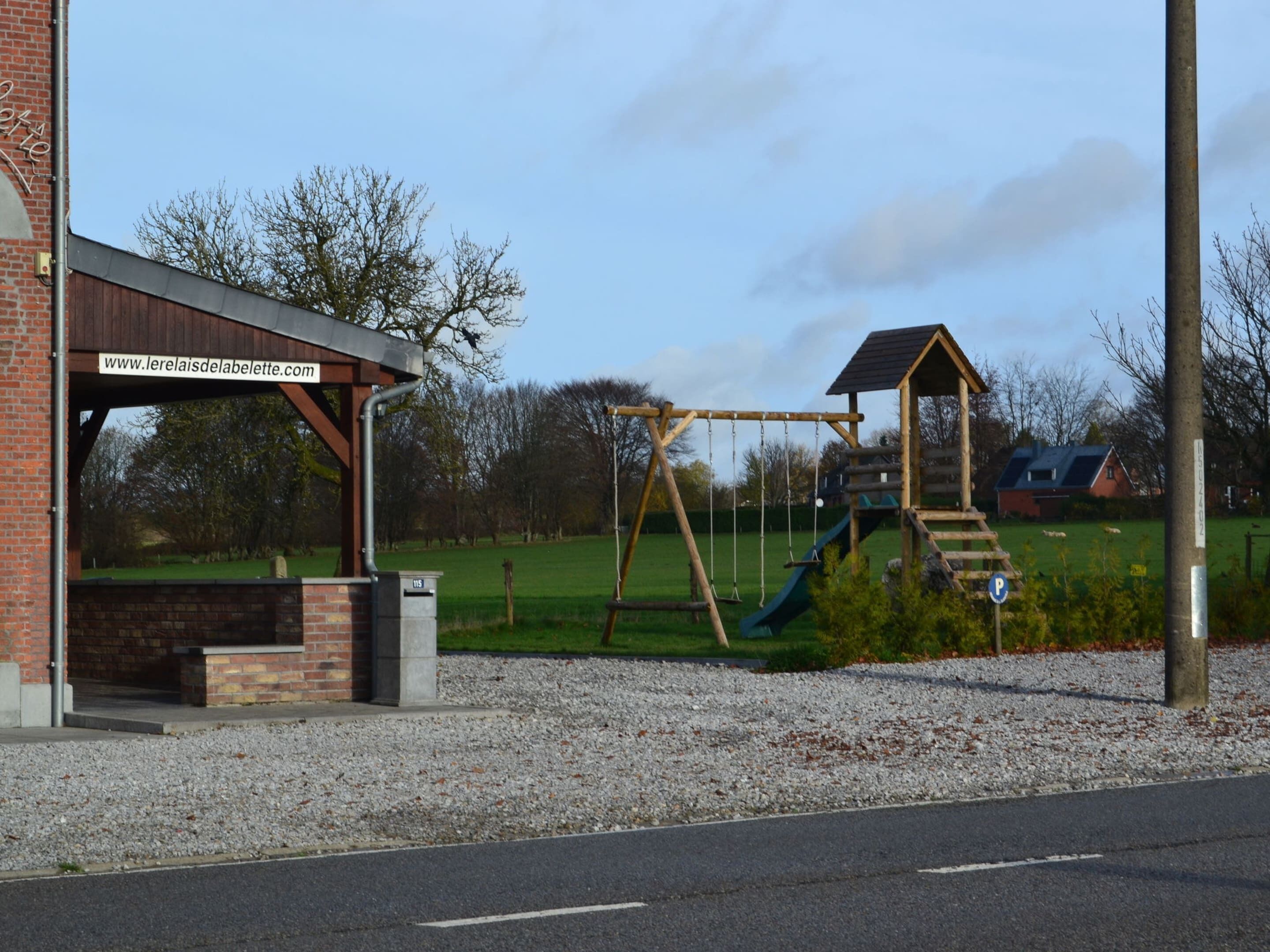 Ein Spielplatz mit Schaukeln und einer Rutsche, ein Backsteinhaus mit einer Website-Adresse und eine Kiesfläche im Vordergrund.