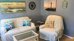 Una habitación con un sofá y un sillón blancos, una mesa de centro de mimbre y dos cuadros en la pared que muestran una playa y un faro.