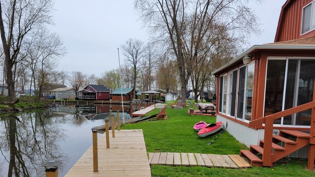 Un muelle de madera que conduce a una superficie acuática tranquila, con un área verde y un edificio rojo con ventanas grandes cerca.