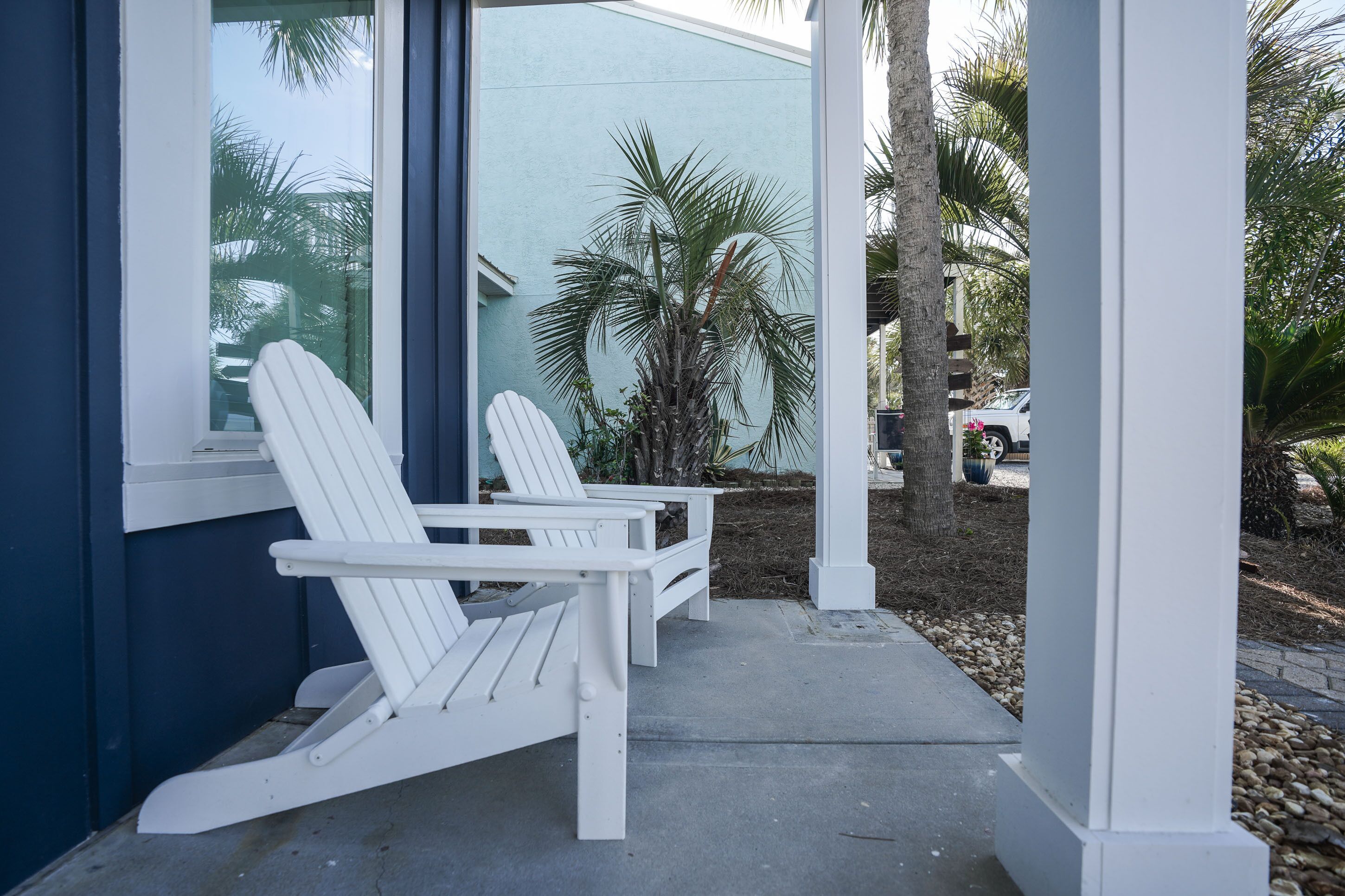 Een overdekt terras met twee witte Adirondack-stoelen en een palmboom op de achtergrond.