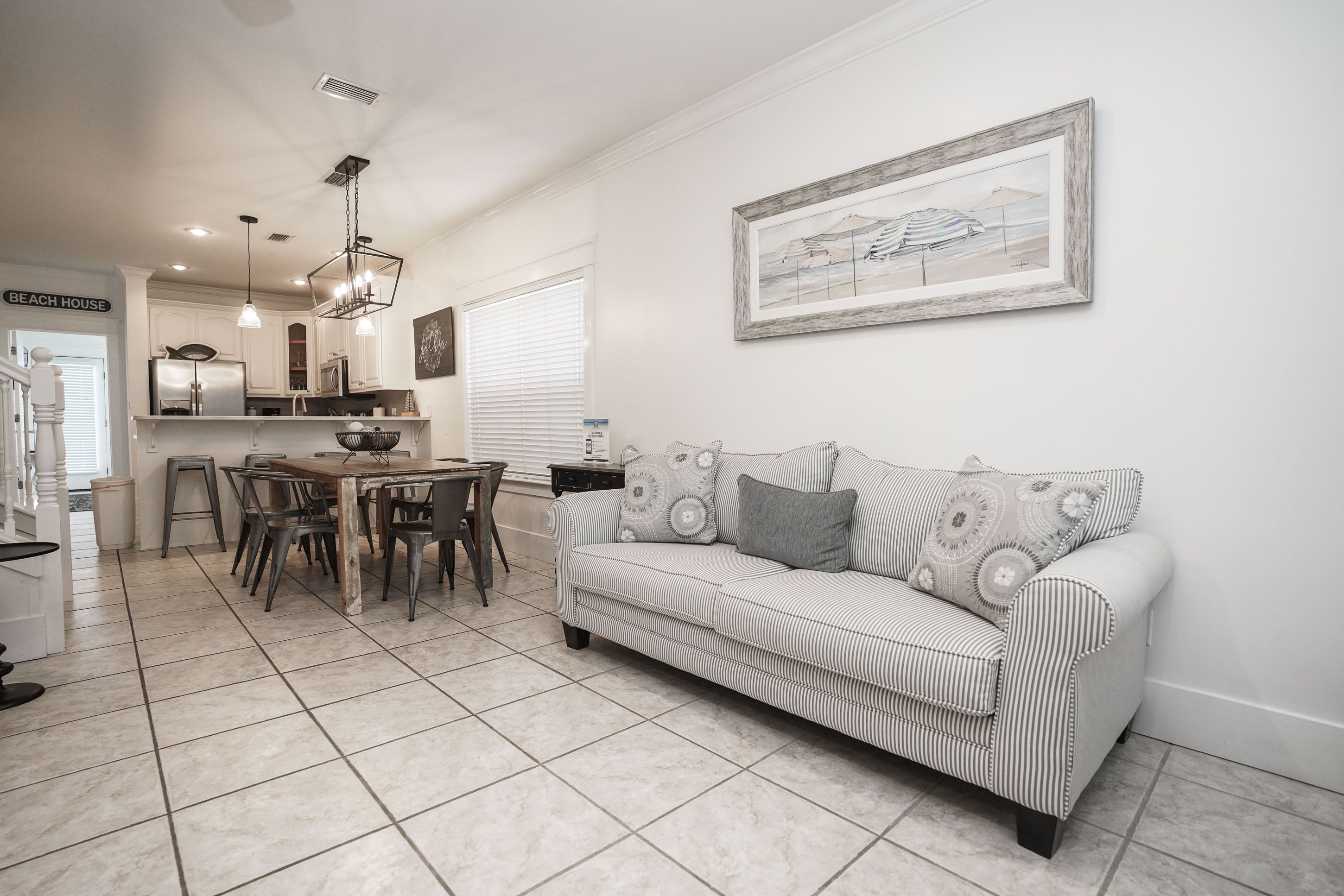 A modern kitchen with white cabinets, a dining area with a striped sofa and patterned cushions, and a framed beach scene on the wall.