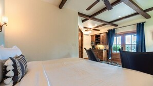 A bedroom with a large bed, a ceiling fan, wooden beams, a window with curtains, and a view of the kitchen. - The Bodega w/personal hot tub & fireplace (Fredericksburg)
