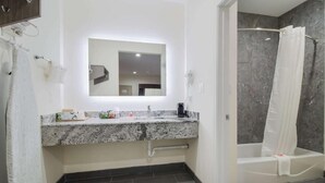 A modern bathroom with a granite countertop, a large mirror, a shower with a curtain, and a towel rack. - REGENCY INN AND SUITES (Humble)