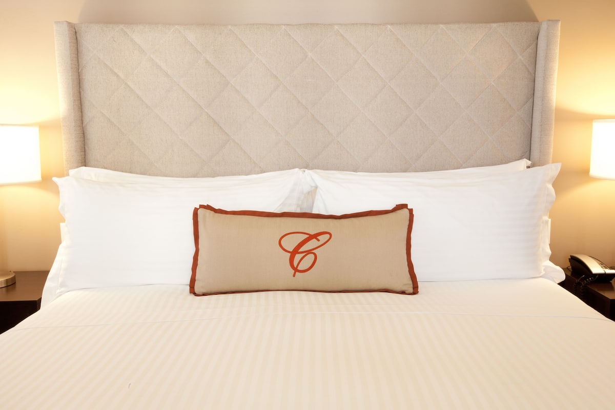 Ein ordentlich bezogenes Bett mit einem gepolsterten Kopfteil, weißer Bettwäsche und einem einzelnen dekorativen Kopfkissen mit Monogramm.
