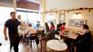Ein Restaurant mit Holztischen und -stühlen, ein Kellner geht vorbei, und an den Wänden befinden sich Regale mit Flaschen und gerahmten Bildern.
