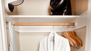 Ein begehbarer Kleiderschrank mit einem weißen Bademantel, der auf einem Gestell hängt, einer Kleiderstange aus Holz und einem schwarzen Föhn auf einem Regal.