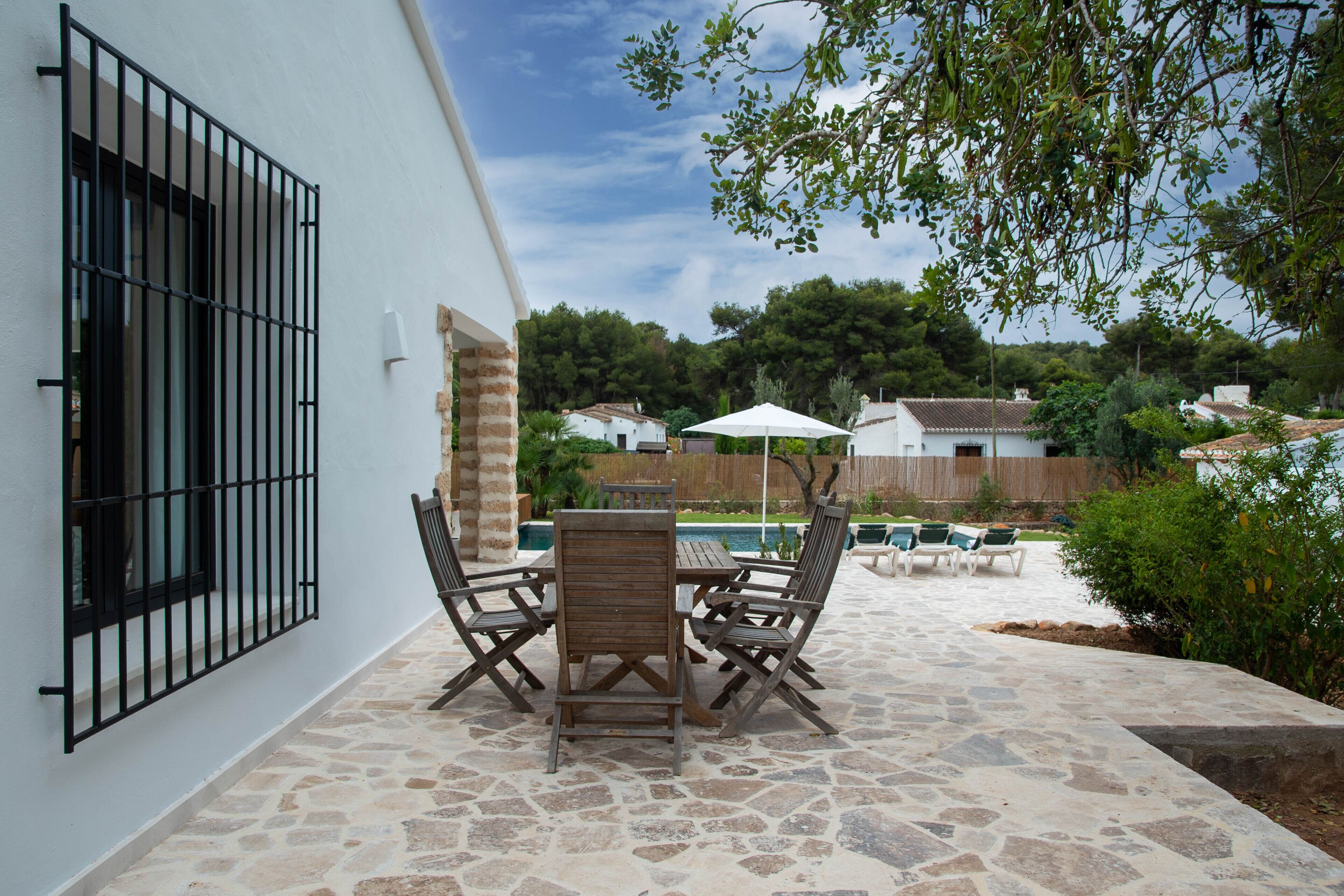 An outdoor patio with stone flooring, a table with chairs, and a pool in the background.