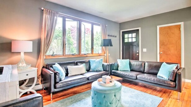 A living room with a sectional sofa, a round ottoman, a side table with a lamp, and a window with curtains. - Fully stocked vacation home with large private yard, decks and BBQ gas grill & 1 mile to Narragansett Beach ! (Narragansett)