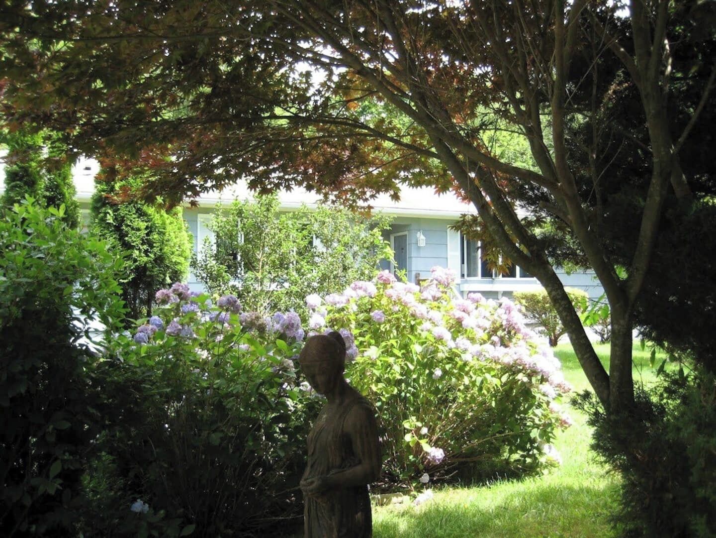 Un jardín con una estatua, abundante vegetación y una casa al fondo.