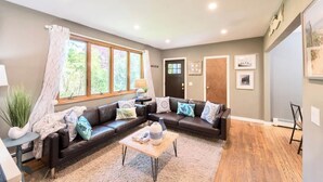 A modern living room with a sectional sofa, a coffee table, and a large window with curtains. - Fully stocked vacation home with large private yard, decks and BBQ gas grill & 1 mile to Narragansett Beach ! (Narragansett)