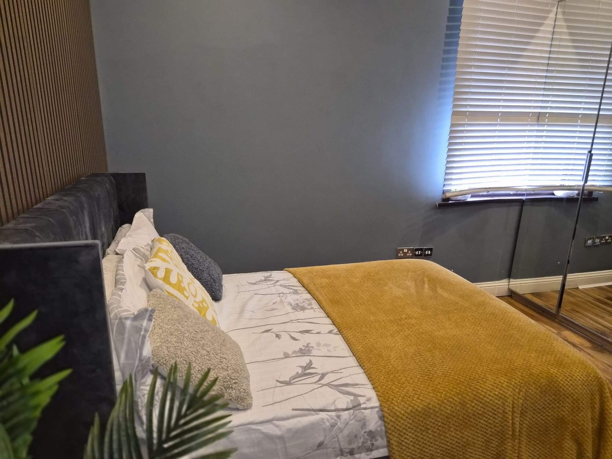 A bedroom with a bed, a bedside table, a window with blinds, and a plant.