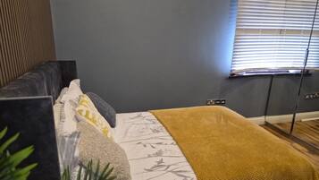 A bedroom with a bed, a bedside table, a window with blinds, and a plant.