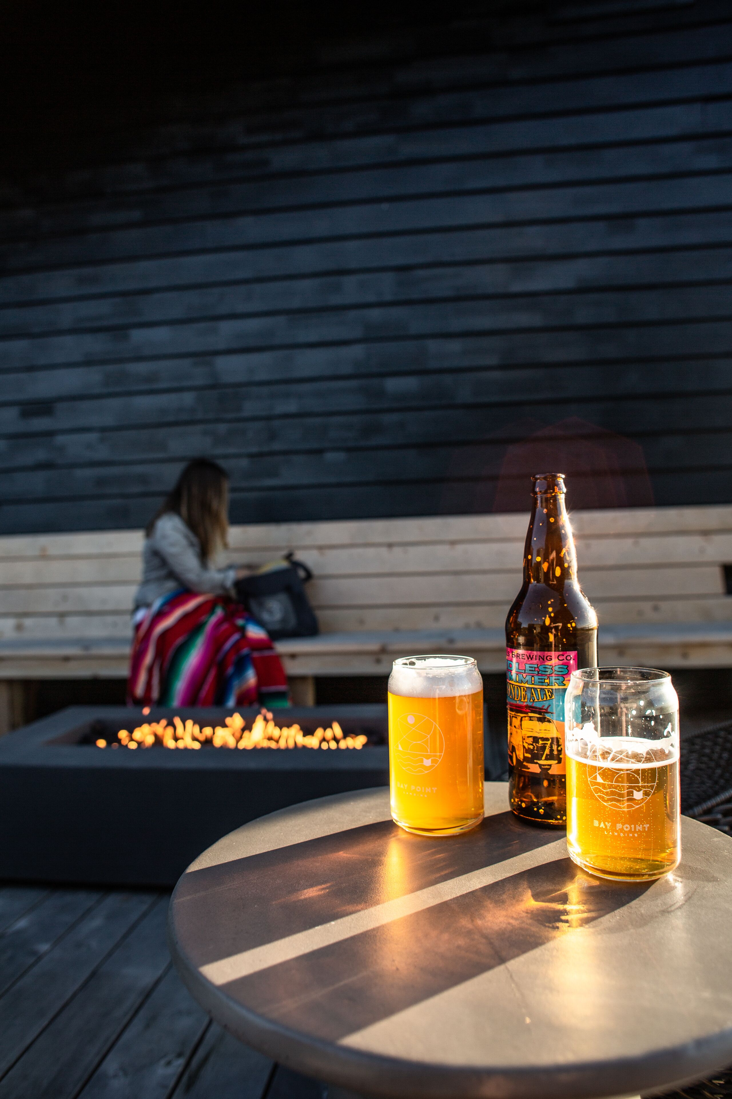 A table with two beer glasses and a bottle, a wooden bench, and a fire pit in the background.