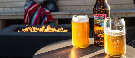 A table with two beer glasses and a bottle, a wooden bench, and a fire pit in the background.